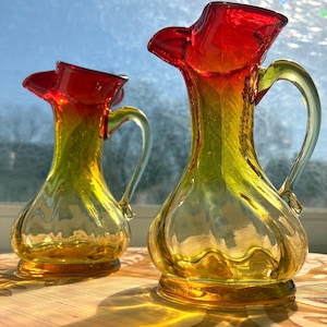 May include: Two vintage glass pitchers with a vibrant color gradient. The rims are a deep red, transitioning to yellow and gold. Each pitcher has a clear handle and sits on a round base. The pitchers are displayed on a wooden surface.