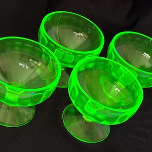 May include: Four vintage green glass dessert bowls. Each bowl has a round base, a stem, and a rounded bowl with a textured pattern around the rim. The bowls are a bright, vibrant green color, and are arranged on a dark surface.