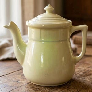 May include: A pale yellow ceramic teapot with a lid and handle. The teapot has a spout and a rounded body. The teapot is sitting on a wooden surface. The teapot is a vintage style.