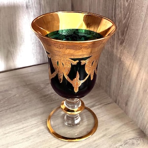 May include: A dark green glass goblet featuring an ornate gold rim and intricate gold filigree patterns around the bowl. The stem is clear glass with a gold-accented base, and the interior of the bowl shows a deep green hue with gold trim.