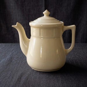 May include: A cream-colored ceramic teapot with a glossy finish, featuring a rounded body, a curved spout, a sturdy handle, and a matching lid with a small finial knob. The upper section of the teapot has a subtle vertical ribbed texture.