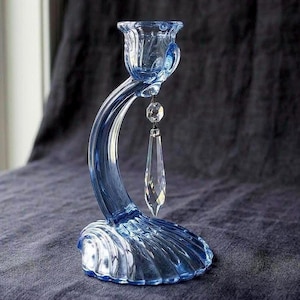 May include: A light blue glass candle holder featuring a curved stem and a base shaped like a seashell. The top cup holds a single clear crystal pendant that hangs from a small metal ring, reflecting light through its faceted, elongated shape.
