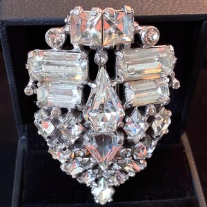 May include: A vintage silver-toned brooch with a dazzling array of clear, faceted crystals. The brooch features a geometric design with rectangular and diamond-shaped stones, creating a sparkling, eye-catching accessory. The brooch is set in a dark box.