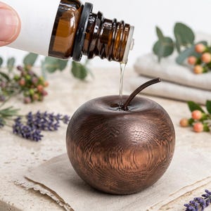 May include: A dark brown wooden apple-shaped diffuser with a visible grain pattern and a thin metal stem. A clear liquid is being poured from an amber glass dropper bottle into the centre of the wooden apple, which sits on a light fabric.