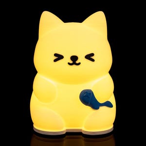 May include: A yellow cat-shaped night light with a blue fish-shaped accessory. The light has a smiling face with closed eyes and is glowing against a black background. The night light is a cute and whimsical decorative item.