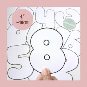 Puede incluir: Un número ocho blanco con contorno negro se sostiene en una mano. El número es parte de un conjunto de números blancos con contornos negros. Un círculo rosa indica que el número mide 10 cm.