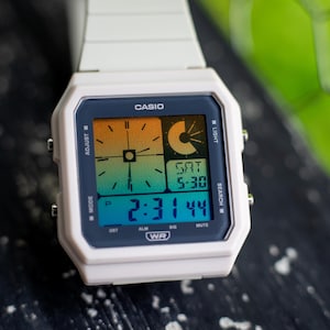 Puede incluir: Un reloj digital Casio blanco con esfera cuadrada y pantalla de degradado multicolor. La pantalla muestra la hora 2:31:44, la fecha SAT 5-30 y varias etiquetas de función que incluyen ADJUST, LIGHT, MODE, SEARCH y WR. El reloj tiene una correa blanca.