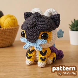 Puede incluir: Una criatura amigurumi de ganchillo con cabeza gris oscuro texturizada, ojos negros grandes y un accesorio en forma de estrella azul claro. El cuerpo es amarillo con rayas negras, detalles blancos y una cola morada hecha de hilo.