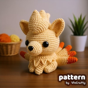 Op de afbeelding: Een handgemaakt gehaakt dier in tinten geel en oranje. Het pluche speelgoed heeft grote zwarte ogen, een puntige snuit en een kroonachtig kenmerk op zijn hoofd. De tekst "pattern by VinCrafty" is zichtbaar.