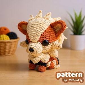 May include: A handmade crochet Arcanine plushie in cream, orange, and black. The plushie has large black eyes and a detailed design, with the text "pattern by VinCrafty" in the lower right corner. A basket of yarn is in the background.