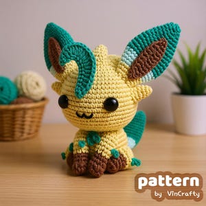 May include: A handmade crochet Leafeon plushie in yellow, green, and brown. The plushie has large, detailed ears, black eyes, and a smiling mouth. The text "pattern by VinCrafty" is visible in the lower right corner. The plushie is sitting on a wooden surface.