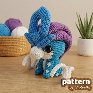Puede incluir: Una figura de criatura tejida a ganchillo con un cuerpo azul brillante, una gran cresta morada, un hocico blanco y detalles blancos. La figura tiene grandes ojos negros y parches blancos en forma de diamante en los costados. El texto pattern by VinCrafty es visible.
