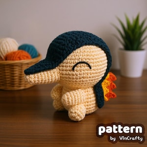 May include: A handmade crochet plush toy in the shape of a cartoon character. The toy is primarily beige with a dark blue head and a small orange and yellow flame detail. The text "pattern by VinCrafty" is visible.