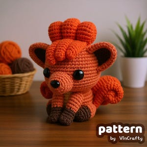 Könnte beinhalten: Ein handgefertigter orangefarbener Häkelfuchs mit schwarzen Augen und einer schwarzen Nase. Der Fuchs hat einen flauschigen Schwanz und Ohren mit braunen Akzenten. Der Text "pattern by VinCrafty" befindet sich in der unteren rechten Ecke. Ein Korb mit Garn ist im Hintergrund.
