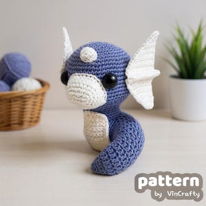 May include: A handmade crochet plush toy in the shape of a sea creature, primarily blue with white accents. It has large black eyes and white fins. The text "pattern by VinCrafty" is visible in the lower right corner.