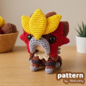 Puede incluir: Una criatura amigurumi de ganchillo con cuerpo marrón, una cresta amarilla brillante y un detalle facial rojo en forma de estrella. Tiene grandes ojos negros, un hocico gris y patas gris claro. El texto "pattern by VinCrafty" es visible.