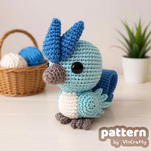 May include: A handmade crochet bird in shades of blue, white, and gray, with large black eyes. The bird has a textured appearance and is positioned in front of a basket with yarn balls. The text "pattern by VinCrafty" is visible.