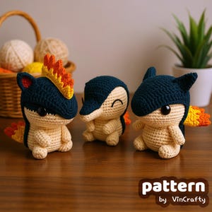 Peut inclure: Trois figurines Cyndaquil faites au crochet. Chacune a une tête bleu marine, un corps crème et une queue en forme de flamme. Les figurines sont posées sur une surface en bois. Le texte "pattern by VinCrafty" est en bas à droite.