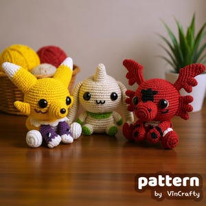 May include: Three handmade crochet figures in yellow, off-white, and red. The yellow figure has fox-like ears and purple accents. The off-white figure has a green collar, and the red figure has wings. The figures are on a wooden surface.