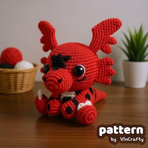 May include: A handmade red crochet dragon with black accents, large black eyes, and small wings. The dragon is sitting on a wooden surface. A basket with red and white yarn is in the background. The text "pattern by VinCrafty" is in the lower right corner.