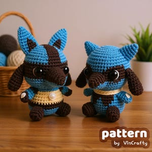 以下が含まれることがあります： 大きな黒い目を持つ青と茶色の生き物の手作りのかぎ針編み人形が2つ。人形は木製の表面に座っています。人形の胸には黄色と白のディテールがあります。「pattern by VinCrafty」というテキストが右下にあります。