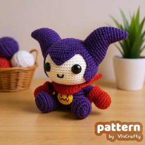Puede incluir: Muñeca de ganchillo hecha a mano en morado, rojo, blanco y amarillo. La muñeca tiene orejas grandes, una bufanda roja y un emblema amarillo. Está sobre una superficie de madera. El texto "pattern by VinCrafty" es visible.