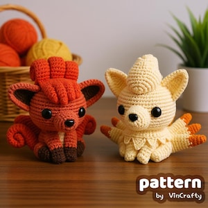 May include: Two crocheted fox-like figures, one orange and one yellow, sit side-by-side. The orange figure has dark brown accents and the yellow figure has orange accents. The text "pattern by VinCrafty" is in the lower right corner.