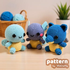 May include: Three handmade crochet turtle toys in blue, purple, and blue. Each turtle has a yellow belly and black eyes. The toys are sitting on a light brown surface. The text "pattern by VinCrafty" is in the bottom right corner.
