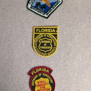 Op de afbeelding: Drie geborduurde patches met tekst en afbeeldingen. De bovenste patch leest "Ranger Florida Park Service" met een natuurscène. De middelste patch zegt "Florida Department of Agriculture & Consumer Services, Division of Forestry". De onderste patch leest "Florida Division of Forestry, Rural Community Fire Protection Cooperator".