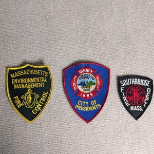 Op de afbeelding: Drie geborduurde patches: Massachusetts Environmental Management Fire Control, een blauwe Quincy Fire Dept patch en een Southbridge Fire Dept Mass patch. Elke patch heeft tekst en gedetailleerde ontwerpen.