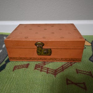 May include: A vintage orange box with a decorative pattern on the lid and a brass clasp. The box has a rectangular shape and is sitting on a green rug.