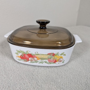 May include: White casserole dish with a brown glass lid and a dark brown knob. The dish features a colorful vegetable design with tomatoes, corn, and mushrooms. The dish has two white handles.