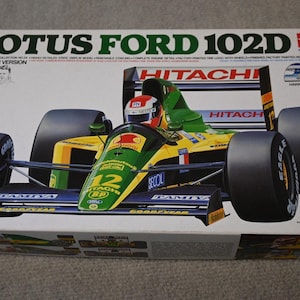 May include: A Tamiya Lotus Ford 102D model kit box. The box features a detailed image of the race car in green, yellow, and blue, with the number 12. The box includes the text "LOTUS FORD 102D" and "HAKKINEN VERSION".
