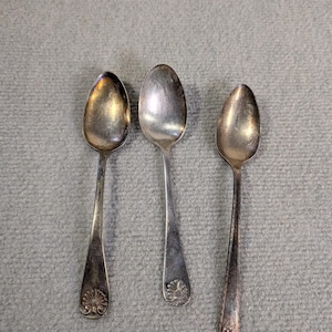 May include: Three antique silver spoons with oval bowls and long handles. The spoons have decorative details at the base of the handles. The spoons are arranged on a gray surface.
