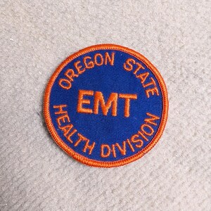 Puede incluir: Un parche circular con un centro azul y un borde naranja. El texto "OREGON STATE HEALTH DIVISION" está arqueado en la parte superior e inferior. La palabra "EMT" está en el centro.