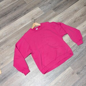 Op de afbeelding: Een felroze sweatshirt met een ronde hals en lange mouwen, gepresenteerd op een houten hanger. De sweatshirt is effen van kleur en lijkt van een zacht materiaal te zijn. Het staat tegen een grijze achtergrond met houtnerf.