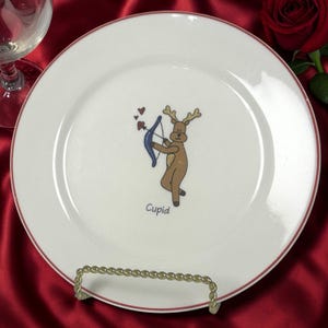 May include: White ceramic plate with a red rim, featuring a cartoon reindeer holding a blue bow and arrow, with the word "Cupid" below. The plate is on a gold-colored metal stand.