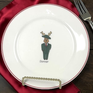 May include: White ceramic plate with a red rim and a cartoon illustration of a reindeer named Donner wearing a green suit and hat. The plate is on a gold-colored stand.