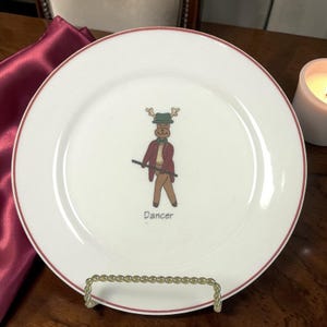 May include: White ceramic plate with a red rim, featuring a cartoon reindeer dressed as a dancer. The reindeer wears a green hat, a burgundy jacket, and holds a cane. The word "Dancer" is printed below the image.
