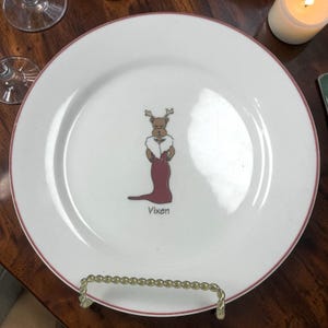 May include: White ceramic plate with a red rim and a gold-colored metal stand. The plate features a cartoon illustration of a reindeer wearing a fur stole and a red dress, with the word "Vixen" below.