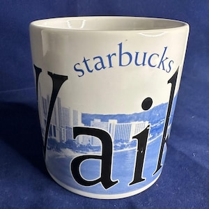 May include: White ceramic Starbucks mug featuring the word "Waikiki" in black, with a blue and white illustration of buildings and the ocean. The word "Starbucks" is in blue. The mug has a cylindrical shape.