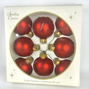 May include: A box of eight red glass ornaments. Each ornament is round with a gold cap and a green wire loop. The box is white with a gold circular insert holding the ornaments. The text "Sparkling Creations" is printed on the box.