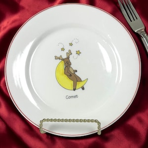 May include: White ceramic plate with a red rim, featuring a cartoon reindeer sitting on a yellow crescent moon. The plate has the word "Comet" written below the moon. Three yellow stars are above the reindeer.