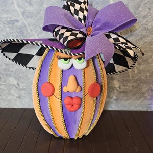 Pu&ograve; includere: Una figura decorativa a forma di zucca con corpo a righe viola e arancioni e dettagli in corda dorata. La figura presenta un volto con guance rosse, naso e labbra rosse, sormontato da un cappello da strega a scacchi bianchi e neri e un grande fiocco viola.
