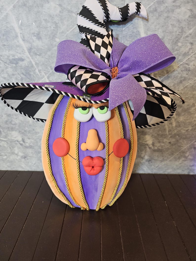 Pu&ograve; includere: Una figura decorativa a forma di zucca con corpo a righe viola e arancioni e dettagli in corda dorata. La figura presenta un volto con guance rosse, naso e labbra rosse, sormontato da un cappello da strega a scacchi bianchi e neri e un grande fiocco viola.