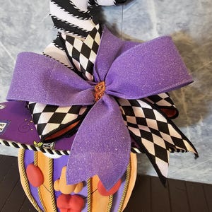 Pu&ograve; includere: Una decorazione da parete raffigurante un cappello da strega a righe bianche e nere con motivo a rombi, ornato da un grande fiocco viola glitterato. Sotto il cappello si trova un volto a forma di zucca con naso, labbra e guance rosse.
