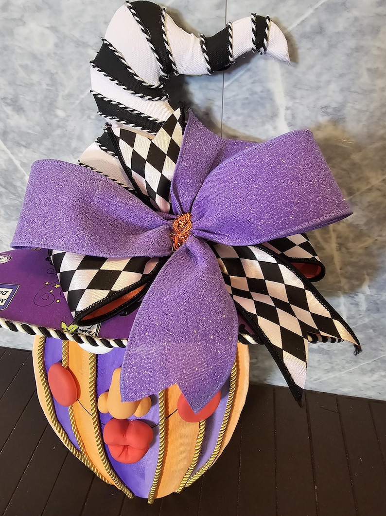 Pu&ograve; includere: Una decorazione da parete raffigurante un cappello da strega a righe bianche e nere con motivo a rombi, ornato da un grande fiocco viola glitterato. Sotto il cappello si trova un volto a forma di zucca con naso, labbra e guance rosse.