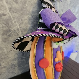 Pu&ograve; includere: Una figura decorativa a forma di zucca con un volto, che indossa un cappello alto e appuntito. La zucca presenta segmenti verticali alternati arancioni e viola separati da un cordoncino dorato. Il cappello mostra un motivo a scacchi bianco e nero, un fiocco viola glitterato e una tesa viola con piccole stampe grafiche. Il volto include guance rosse rotonde, un naso color pesca, labbra rosse e occhi verdi con lunghe ciglia.