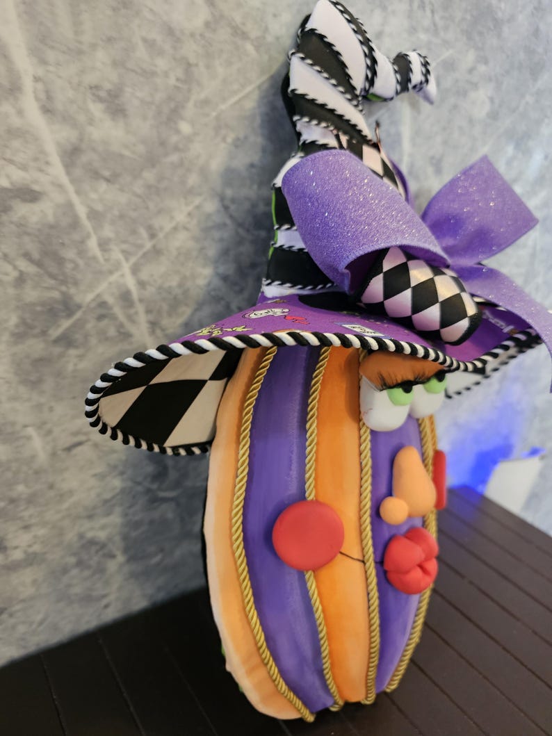 Pu&ograve; includere: Una figura decorativa a forma di zucca con un volto, che indossa un cappello alto e appuntito. La zucca presenta segmenti verticali alternati arancioni e viola separati da un cordoncino dorato. Il cappello mostra un motivo a scacchi bianco e nero, un fiocco viola glitterato e una tesa viola con piccole stampe grafiche. Il volto include guance rosse rotonde, un naso color pesca, labbra rosse e occhi verdi con lunghe ciglia.