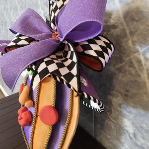 Pu&ograve; includere: Una decorazione da parete con un grande fiocco viola glitterato sovrapposto a un nastro a scacchi bianchi e neri. La base &egrave; una forma di zucca viola e arancione, arricchita da un bordo in corda dorata e piccole forme in argilla rosse.